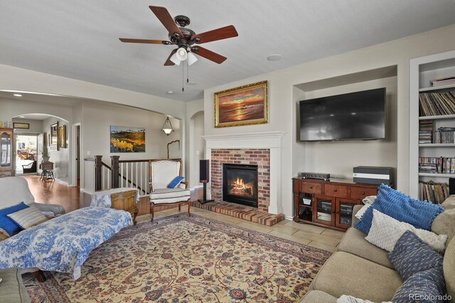 tiled living room with arched walkways, a brick fireplace, and ceiling fan