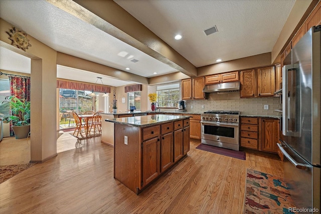 kitchen featuring brown cabinets, dark stone counters, light wood-type flooring, under cabinet range hood, and premium appliances
