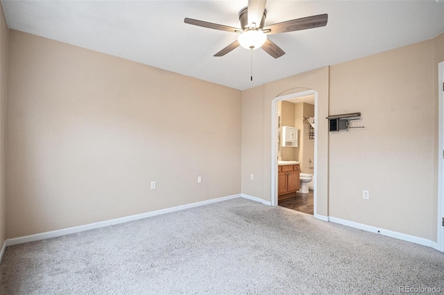 unfurnished bedroom with arched walkways, dark colored carpet, baseboards, ensuite bath, and ceiling fan