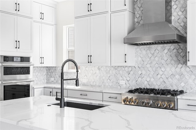 kitchen featuring wall chimney range hood, white cabinets, a sink, stainless steel appliances, and decorative backsplash