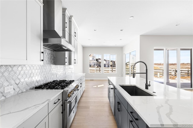 kitchen featuring wall chimney range hood, appliances with stainless steel finishes, a center island with sink, a sink, and light wood-style floors