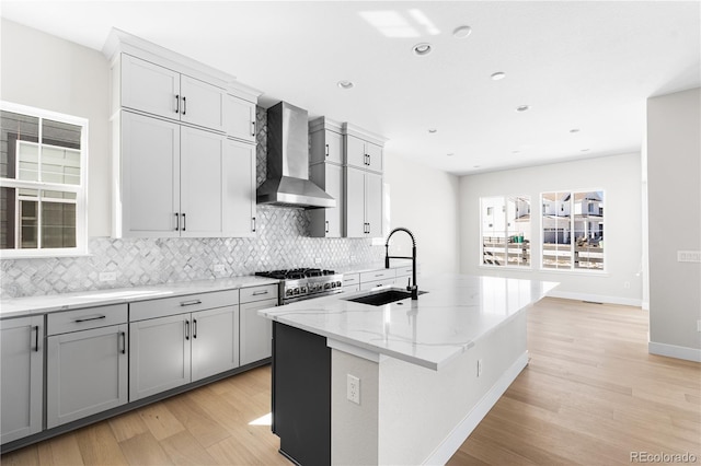 kitchen featuring wall chimney range hood, light stone counters, tasteful backsplash, a sink, and light wood-type flooring