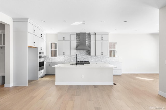 kitchen with decorative backsplash, baseboards, a center island with sink, wall chimney exhaust hood, and light wood finished floors