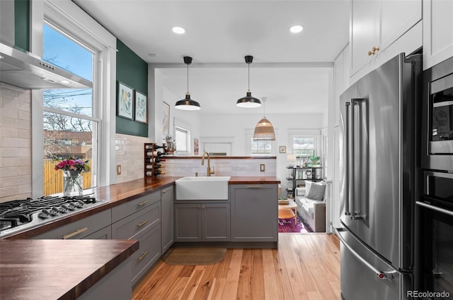 kitchen featuring butcher block countertops, gray cabinetry, hanging light fixtures, stainless steel appliances, and wall chimney range hood