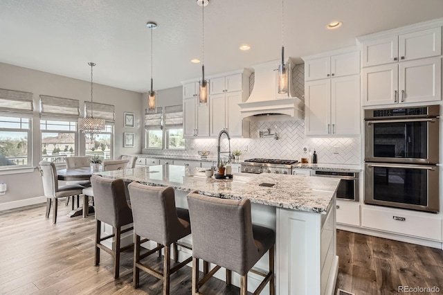 kitchen with light stone countertops, appliances with stainless steel finishes, a kitchen island with sink, premium range hood, and tasteful backsplash