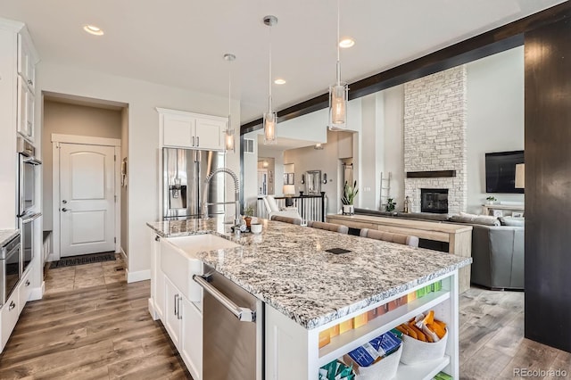 kitchen with open shelves, a kitchen island with sink, a stone fireplace, a sink, and appliances with stainless steel finishes