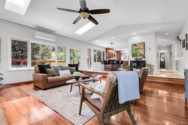 living room with wood finished floors, an AC wall unit, a ceiling fan, lofted ceiling with skylight, and baseboards