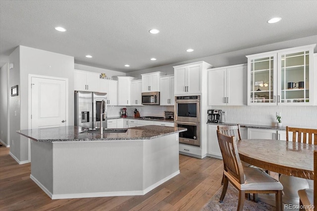 kitchen featuring wood finished floors, appliances with stainless steel finishes, backsplash, dark stone countertops, and a center island with sink