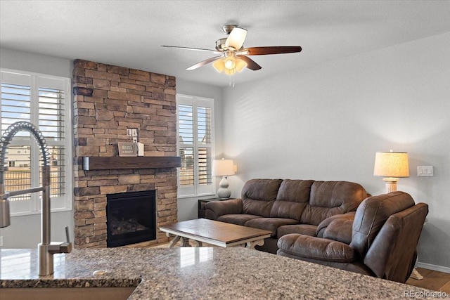 living area featuring a textured ceiling, a stone fireplace, wood finished floors, a ceiling fan, and baseboards