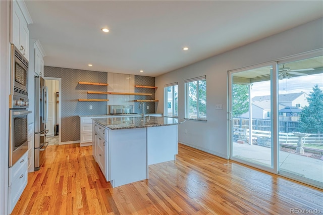 kitchen with stone counters, open shelves, stainless steel appliances, a kitchen island with sink, and white cabinetry