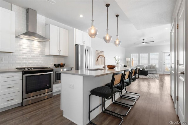 kitchen featuring wall chimney range hood, a sink, stainless steel appliances, dark wood-type flooring, and a kitchen breakfast bar