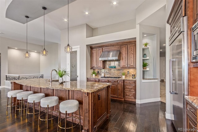 kitchen featuring ventilation hood, hanging light fixtures, decorative backsplash, light stone countertops, and an island with sink