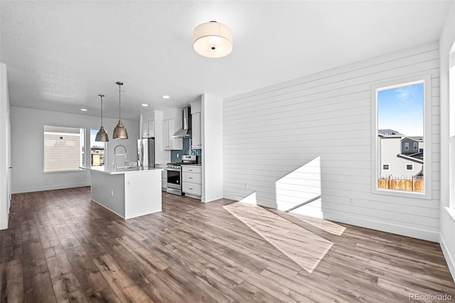 kitchen featuring white cabinetry, decorative light fixtures, a center island with sink, appliances with stainless steel finishes, and wall chimney range hood