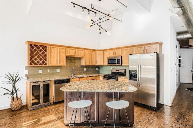 kitchen with beverage cooler, appliances with stainless steel finishes, hardwood / wood-style flooring, a towering ceiling, and a center island