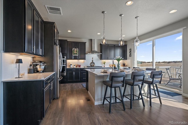 kitchen featuring wall chimney range hood, dark wood-style floors, visible vents, a kitchen breakfast bar, and dark cabinets