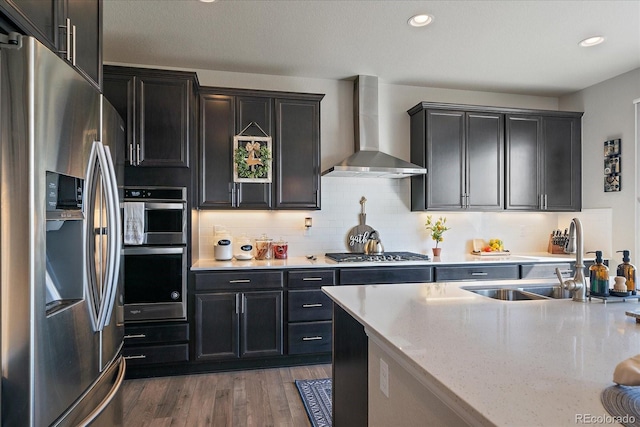 kitchen with wood finished floors, dark cabinetry, appliances with stainless steel finishes, wall chimney range hood, and decorative backsplash