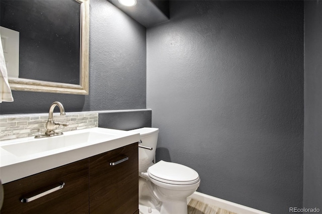 half bathroom featuring baseboards, toilet, decorative backsplash, a textured wall, and vanity