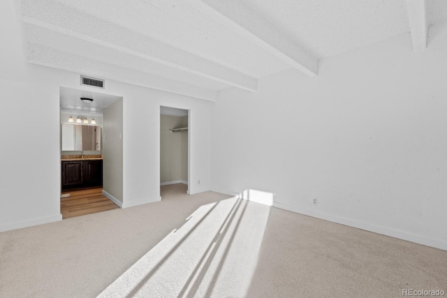 unfurnished bedroom featuring a spacious closet, beamed ceiling, ensuite bathroom, light carpet, and a closet