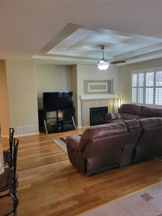 living room featuring ceiling fan, a fireplace, a raised ceiling, and light wood-type flooring