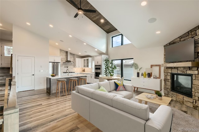 living area with ceiling fan, high vaulted ceiling, a stone fireplace, and light wood-type flooring