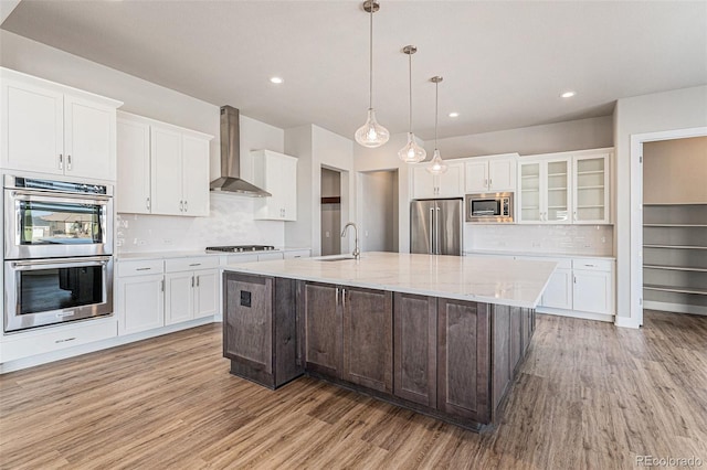 kitchen featuring sink, white cabinets, a kitchen island with sink, stainless steel appliances, and wall chimney range hood