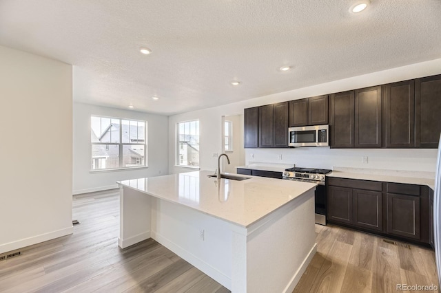 kitchen featuring appliances with stainless steel finishes, a sink, light wood finished floors, a textured ceiling, and a center island with sink