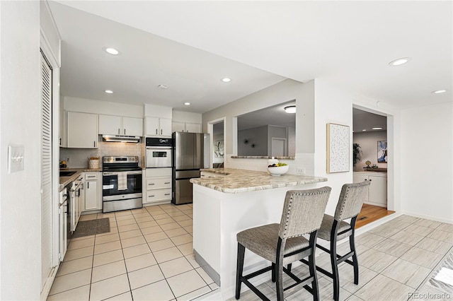 kitchen featuring stainless steel appliances, a breakfast bar area, decorative backsplash, light tile patterned flooring, and under cabinet range hood