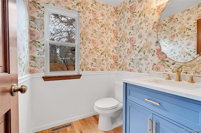 half bathroom with visible vents, toilet, wood finished floors, wainscoting, and wallpapered walls