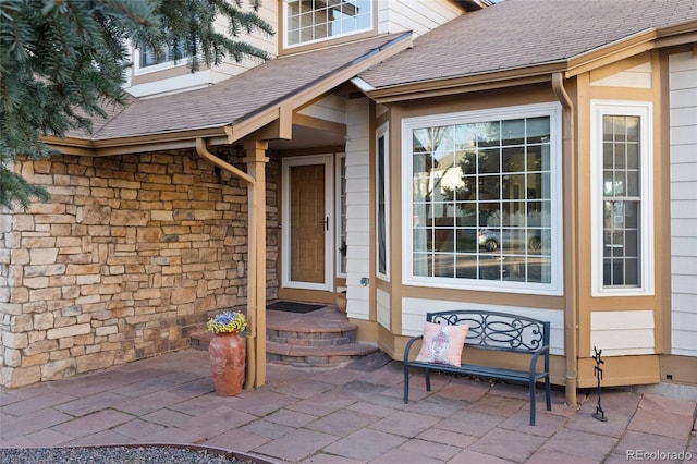 entrance to property with stone siding, roof with shingles, and a patio