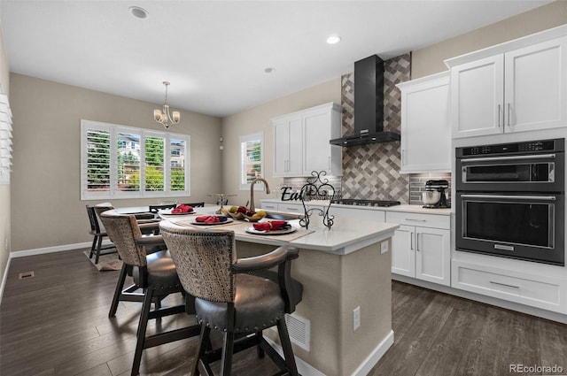 kitchen featuring wall chimney exhaust hood, stainless steel appliances, an island with sink, and white cabinetry