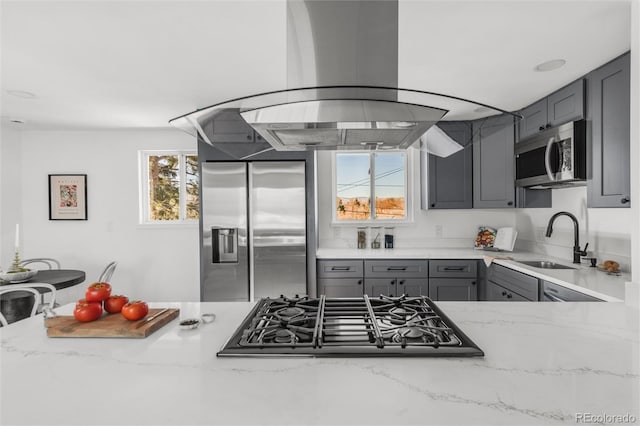 kitchen with sink, light stone counters, appliances with stainless steel finishes, gray cabinets, and island exhaust hood