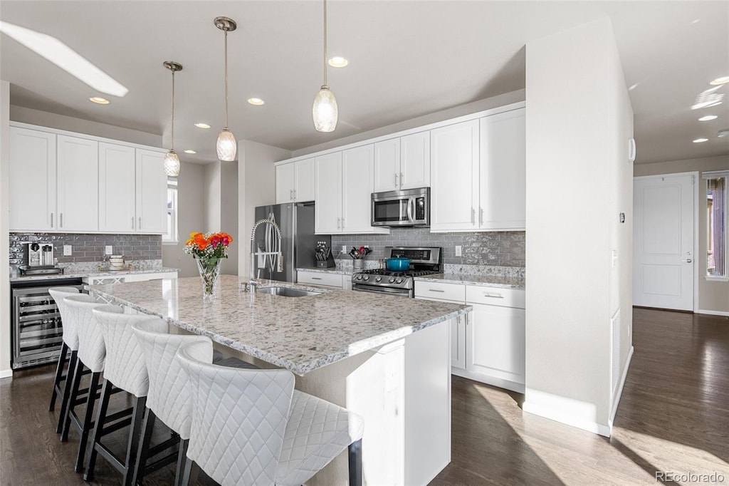 kitchen with beverage cooler, appliances with stainless steel finishes, hanging light fixtures, an island with sink, and white cabinets