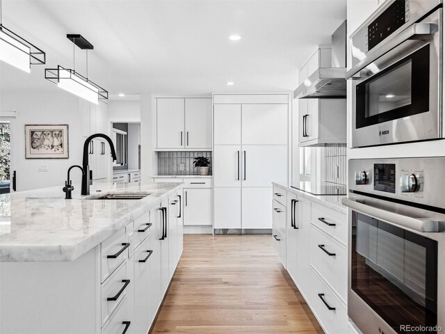 kitchen featuring wall chimney range hood, an island with sink, a sink, and stainless steel oven