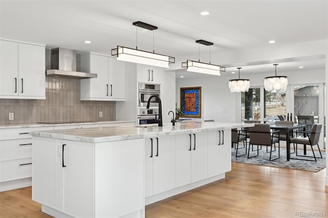 kitchen featuring wall chimney range hood, white cabinetry, a large island with sink, and pendant lighting