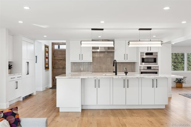 kitchen featuring stainless steel double oven, white cabinetry, light stone countertops, a center island with sink, and pendant lighting