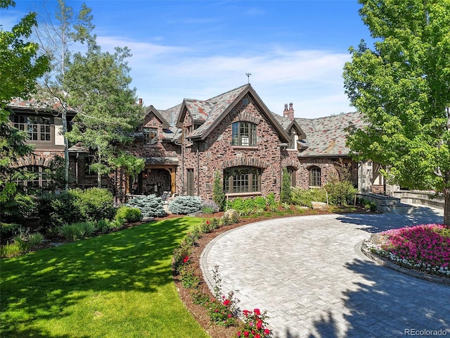 english style home with a high end roof, a chimney, curved driveway, a front yard, and brick siding