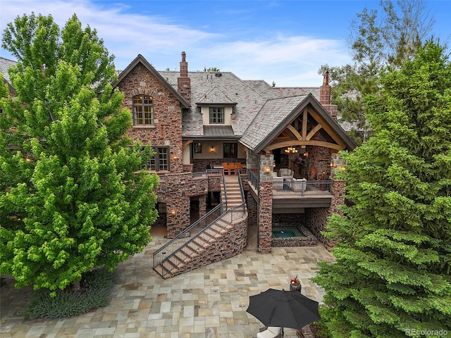 back of property with a high end roof, stairway, stone siding, a chimney, and a patio
