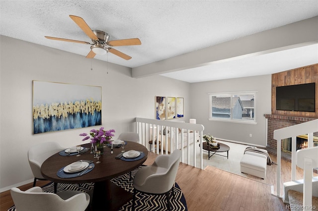 dining area featuring beamed ceiling, ceiling fan, wood-type flooring, a fireplace, and a textured ceiling