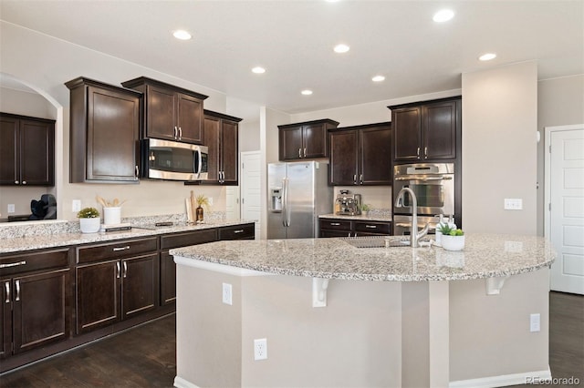 kitchen with light stone counters, a kitchen island with sink, stainless steel appliances, dark wood-type flooring, and dark brown cabinetry