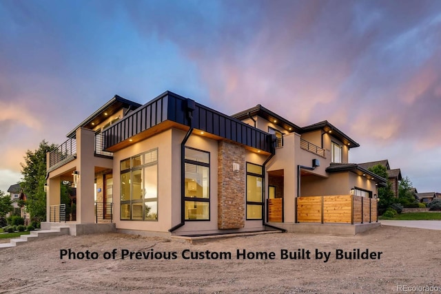 back of property at dusk featuring a balcony, stone siding, board and batten siding, and stucco siding