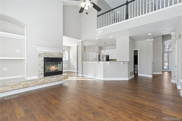 unfurnished living room featuring a stone fireplace, dark hardwood / wood-style flooring, ceiling fan, decorative columns, and a high ceiling