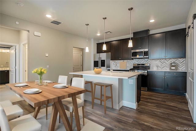 kitchen with appliances with stainless steel finishes, a kitchen island with sink, sink, dark wood-type flooring, and tasteful backsplash