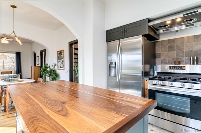 kitchen featuring arched walkways, wooden counters, appliances with stainless steel finishes, wall chimney exhaust hood, and tasteful backsplash