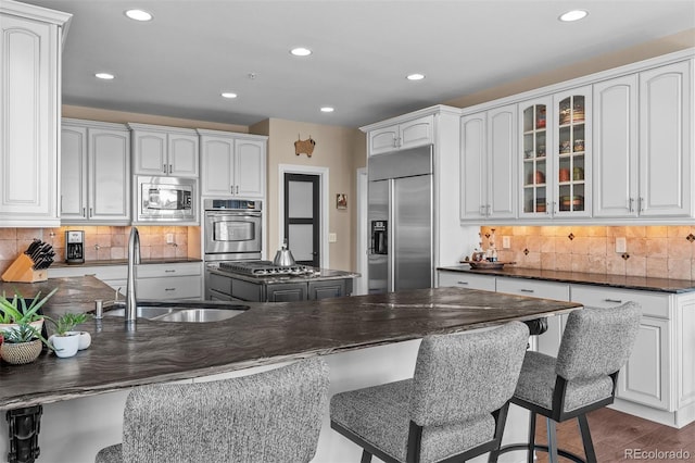 kitchen with built in appliances, a sink, white cabinetry, a kitchen bar, and glass insert cabinets