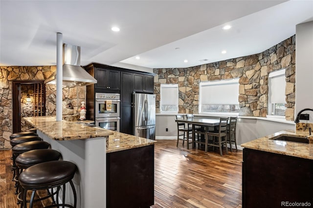 kitchen featuring dark wood-style floors, island exhaust hood, appliances with stainless steel finishes, a sink, and light stone countertops