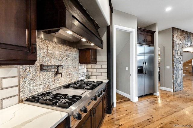 kitchen featuring dark brown cabinetry, arched walkways, wall chimney exhaust hood, appliances with stainless steel finishes, and backsplash