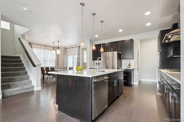 kitchen with dark hardwood / wood-style floors, hanging light fixtures, a kitchen island with sink, stainless steel appliances, and wall chimney range hood