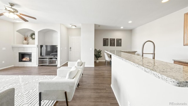 kitchen featuring a tile fireplace, dark wood-type flooring, ceiling fan, and light stone counters