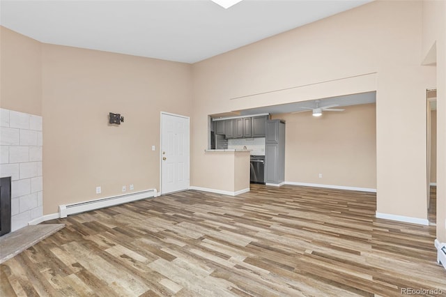 unfurnished living room featuring a tile fireplace, a baseboard radiator, a high ceiling, ceiling fan, and light hardwood / wood-style floors