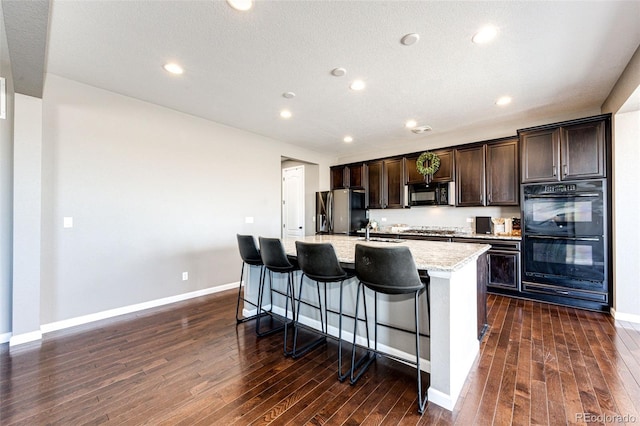 kitchen with dark wood-style flooring, a kitchen island with sink, dark brown cabinets, black appliances, and a sink